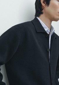 Dunkle Strickjacke mit Kragen, ausgestattet mit zwei Knöpfen und einer strukturierten Oberfläche, getragen über einem weißen Hemd mit Kragen.