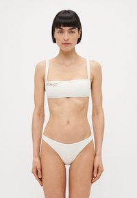 Balts bikini komplekts ar bandeau stilu augšu, kas rotāts ar viļņainām siksnām un smalku ziedu izšuvumu, saskaņots ar zemās jostasvietas apakšdaļu.