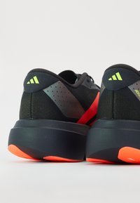 Des chaussures de sport noires avec une tige en maille texturée, présentant des accents vert fluo, des rayures rouges et une épaisse semelle noire avec une semelle extérieure orange.