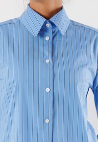 Camicia di jeans azzurro chiaro realizzata in tessuto morbido, con strisce bianche verticali, colletto a punta e bottoni bianchi.