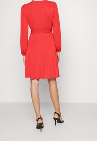 Červené šaty po kolena s dlouhými nabíranými rukávy a podepnutým páskem v pase. Hladká tkanina a silueta ve tvaru písmene A. Nosené s černými sandály na vysokém podpatku.