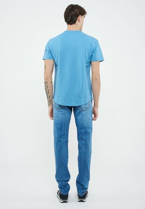 Uomo in piedi di spalle, indossa una maglietta azzurro chiaro, jeans blu, sneakers nere, con un tatuaggio floreale sull'avambraccio sinistro.