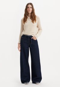 Krmfärgad öppenstickad tröja med ribbade muddar kombinerad med högmidjade, vida mörka denimjeans. Silverskor kompletterar outfiten.