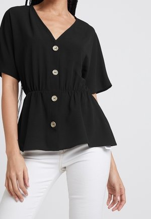 Kobieta ubrana w czarną bluzkę z dekoltem w serek, krótkim rękawem, z czterema guzikami i marszczonym paskiem w talii, zestawioną z białymi spodniami na jednolitym tle.