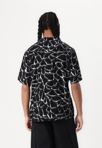 Krátký rukáv košile z černé tkaniny s bílým abstraktním vzorem. Volný střih s špičatým límcem a rovnym lemem.