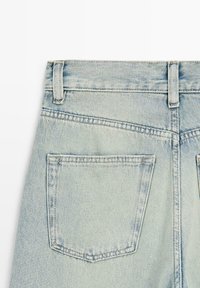 Jean en denim bleu clair vu de dos, montrant les passants de ceinture et une seule poche plaquée carrée sur le côté gauche.