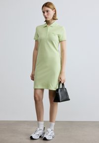 Robe polo vert clair à manches courtes, patte de boutonnage et logo brodé. Associée à un sac à main noir et des baskets blanches. Texture lisse.