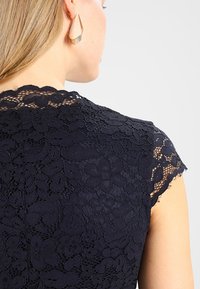 Mörkblå spetsklänning med blommönster, korta kapärmar och en scalloped halsringning. Texturerad design ger djup och detalj.