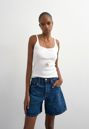 Jeune femme debout, une main dans la poche, portant un débardeur blanc, un short en jean bleu et un long collier en argent avec un pendentif.