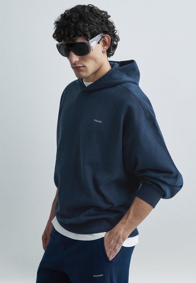 Marineblauer übergroßer Hoodie mit Kordelzug, versehen mit einem kleinen weißen Logo auf der Brust. Kombiniert mit passenden marineblauen Hosen. Strukturierter Stoff.