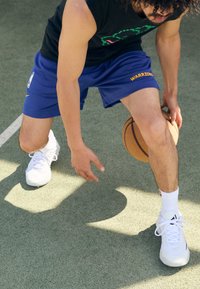 Kosárlabdázó kék rövidnadrágban, amelyen a "WARRIORS" logó látható, fekete ujjatlan pólóban és fehér sportcipőben kosárlabdát cselez egy pályán.