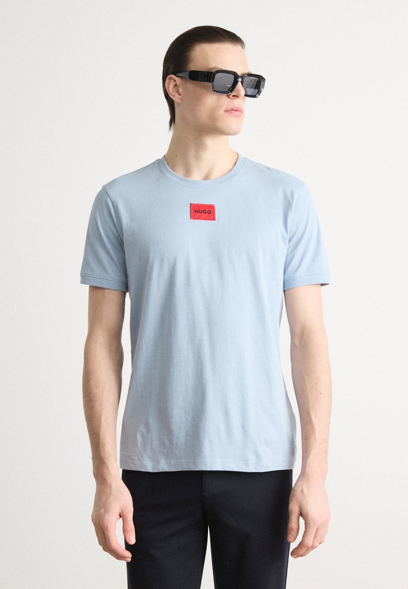 Tricou bleu deschis cu mânecă scurtă, având o etichetă rectangulară roșie cu textul "HUGO." Modelul poartă ochelari de soare pătrați negri, cu un design minimalist.