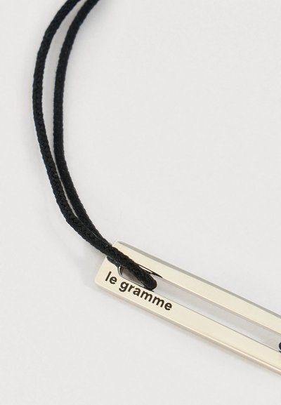 Ezüst téglalap alakú medál, "le gramme" felirattal, fekete fonott zsinórra fűzve, fehér háttér előtt.