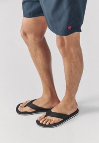 Tongs noires avec des brides texturées et des semelles blanches, portées sur des pieds nus. Le fond est une surface claire unie.