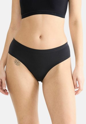 Frau in einem nahtlosen schwarzen Unterwäsche-Set, das den Bauch zeigt, mit einem kleinen lächelnden Blumentattoo auf der linken Hüfte.