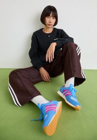 Blå och rosa sneakers med en texturerad ovandel, vita detaljer och gummisula. Modellen bär bruna byxor med mönster och en svart sweatshirt.
