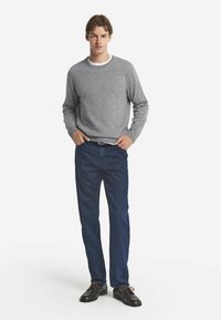 Grau melierter Rundhals-Pullover aus Strickstoff, kombiniert mit dunkelblauen gerade geschnittenen Jeans. Das Modell steht mit den Händen in den Taschen und trägt schwarze Schuhe.