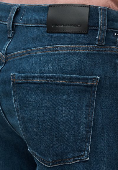 Σκούρα μπλε τζιν με υφή, με ορατή πίσω τσέπη και μαύρη δερμάτινη ετικέτα στη μέση με λογότυπο. Λεπτομέρειες με ραφές αντίθετου χρώματος.