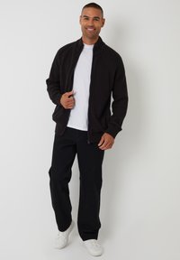 Svart stickad bomberjacka med hel dragkedja, spridd krage, ribbade ärmar, bärs över en vit t-shirt och matchas med svarta byxor och vita sneakers.