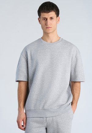 Ung mand med kort mørkt hår iført en simpel lysegrå kortærmet sweatshirt og matchende bukser, stående mod en lys baggrund.