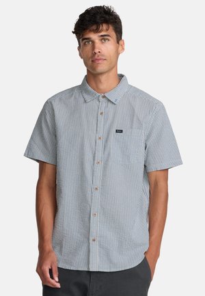 Homme portant une chemise à manches courtes rayée gris et blanc avec une poche poitrine, debout devant un fond uni.