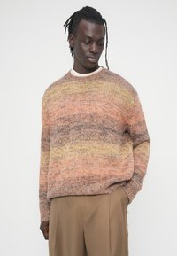 Πλεκτό πουλόβερ σε ζεστές αποχρώσεις πορτοκαλί και μπεζ με άνετη γραμμή, ριμπ μανσέτες και στρογγυλή λαιμόκοψη. Φορεμένο με παντελόνια σε ουδέτερους τόνους.