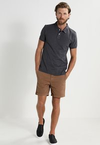 Mørkegrå polo skjorte med to-knappers placket, parret med tan shorts. Modellen har på seg svarte slip-on sko, stående mot en enkel bakgrunn.