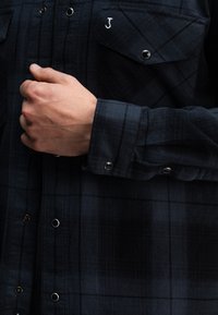 Marine gestreepte flanellen shirt met metalen drukknoopsluitingen, voorzien van twee borstzakken en een klein geborduurd logo op de linkerzak.