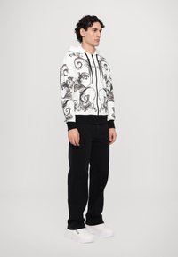 Felpa bianca con motivi floreali neri a spirale, con zip frontale, cordini e polsini neri. Indossata con pantaloni neri e sneakers bianche.