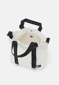 Sac cabas blanc avec poignées noires marqué "Tommy Jeans,", ouverture supérieure montrant une poche intérieure et une fermeture magnétique.