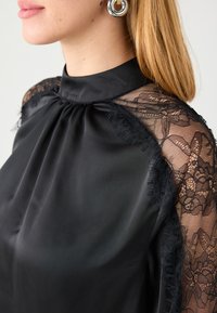 Schwarze Bluse mit hohem Kragen aus Satin und transparenten Spitzenärmeln, verziert mit floralem Muster und ausgefransten Kanten.