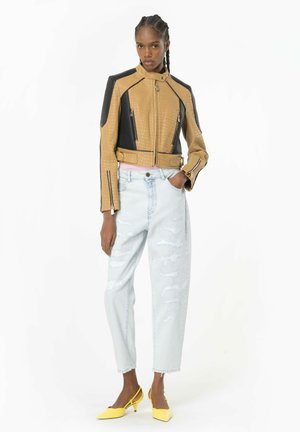 Giacca in pelle beige e nera con texture a coccodrillo e dettagli con zip, abbinata a jeans chiari strappati e tacchi gialli.