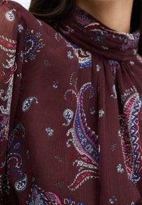 Blouse bordeaux en tissu léger avec un motif paisley bleu, blanc et rose. Le design inclut un col montant et des détails froncés.