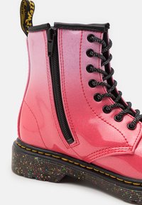 Różowe błyszczące botki za kostkę z bocznym zamkiem błyskawicznym, czarnymi sznurowadłami i nakrapianą platformą. Charakteryzują się błyszczącym materiałem i kontrastującym przeszyciem.