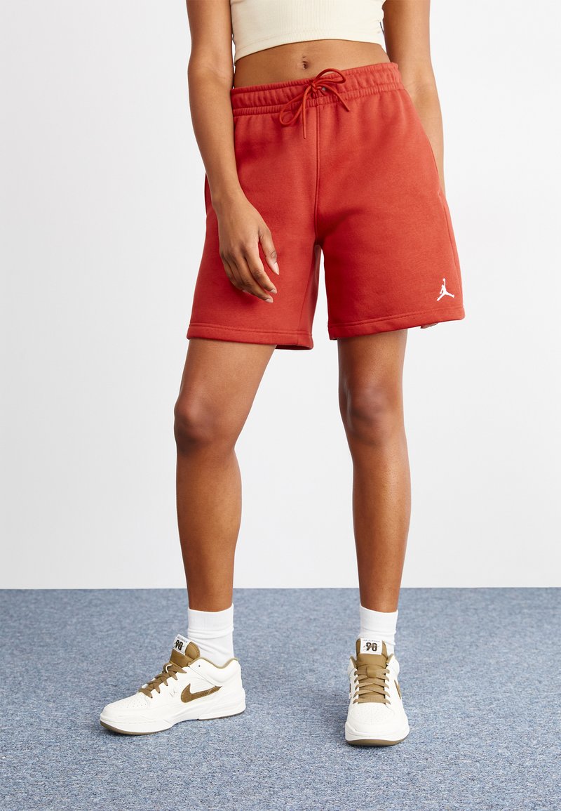 Shorts en coton rouges avec une taille élastique et un cordon de serrage, arborant un logo Nike blanc sur la cuisse gauche. Portés avec des baskets blanches et marron.