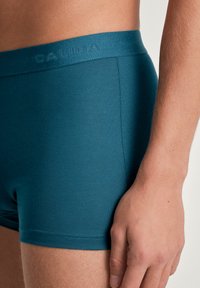 Teal boxershorts van gladde stof met een gestructureerde tailleband waarop "CALIDA" in een subtiele ontwerp staat. Close-up van de pasvorm en beenopening.