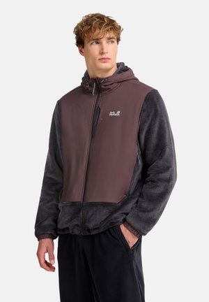 Junger Mann mit lockigem Haar, der eine braune und dunkelgraue Jack Wolfskin Fleecejacke mit Kapuze trägt und die Hand in der Tasche hat.