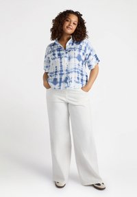 Camicia a maniche corte con bottoni in un motivo tie-dye blu e bianco, abbinata a pantaloni bianchi larghi e scarpe con dettagli blu scuro. Tessuto morbido e vestibilità rilassata.