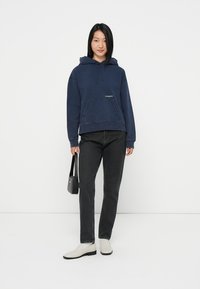 Mørkeblå hettegenser med frontlomme, med en hvit logo, kombinert med svarte jeans og lyse sko. Enkel design.