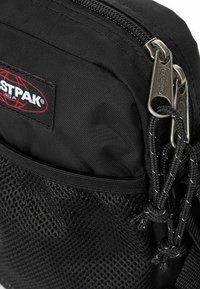 Borsa in nylon nero con tasca in rete, doppie cerniere e etichetta del logo. Presenta una superficie testurizzata e cordini annodati per la chiusura.