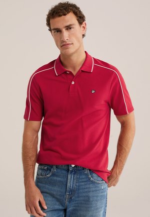 Muž v červenom poloshirte s bielymi lemami a modrých džínsach, stojaci s jednou rukou v vrecku proti jednoliatemu pozadiu.