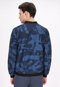 Blauwe bomberjack met een geometrisch patroon in verschillende tinten, voorzien van een zwarte kraag en manchetten, een gladde textuur en een aansluitend ontwerp.