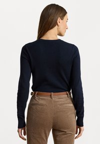 Maglione blu navy a maniche lunghe e collo rotondo abbinato a pantaloni di velluto a coste marrone chiaro e una cintura in pelle marrone, visto da dietro.