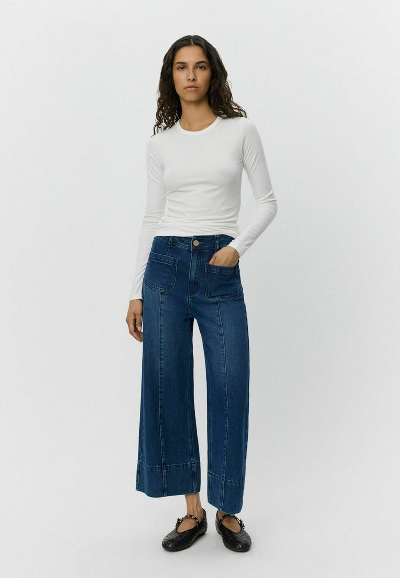 Bijela majica dugih rukava uparena s širokim plavim trapericama koje imaju dva prednja džepa i strukturirani rub. Crne ravne cipele upotpunjuju izgled.