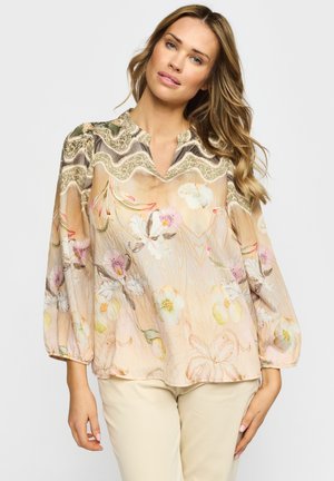 Kvinne med bølgete hår iført en løs, langermet beige bluse med blomstermønster og bølgemønster, kombinert med lyse bukser.