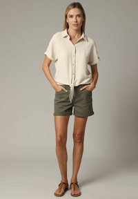 Lichtbeige kortebloesem met knopen aan de voorkant en een gestrikt zoom, gecombineerd met olijfgroene shorts en bruine sandalen. Glad textuur, casual ontwerp.