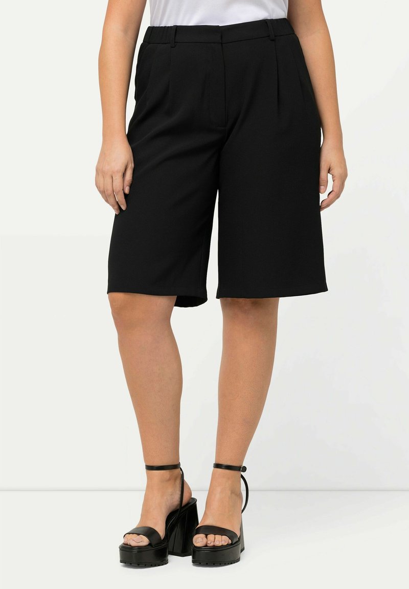 Zwarte op maat gemaakte bermuda shorts met plooien, halverwege de dij, en riemlussen, gecombineerd met chunky zwarte plateausandalen.