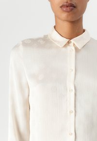 Cream-colored blouse with a button-down collar, featuring a subtle textured pattern and long sleeves. Buttons are clear with a slight sheen.