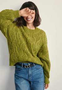 Zielony sweter z dzianiny z warkoczowym wzorem i prążkowanym okrągłym dekoltem, w połączeniu z niebieskimi, wysokim stanem jeansami oraz czarnym paskiem z podwójną klamrą.