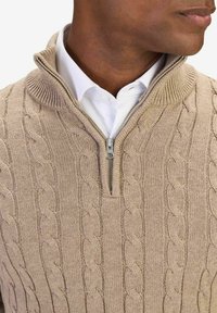 Beige stickad tröja med kvartzip-krag. Har kabelstickat mönster och ribbade kanter. Bärs över en vit skjorta med krage.